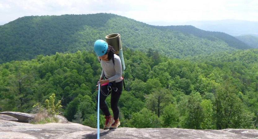 A person wearing safety gear is secured by ropes as they stand near the edge of a cliff, high above a mountainous landscape. 